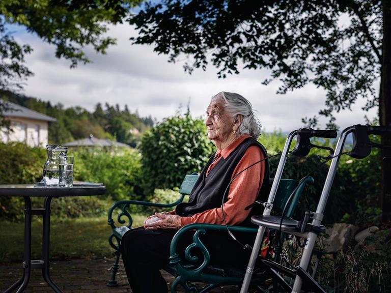 Alte Dame sitzt nachdenklich auf Parkbank, daneben ihr Rollator.