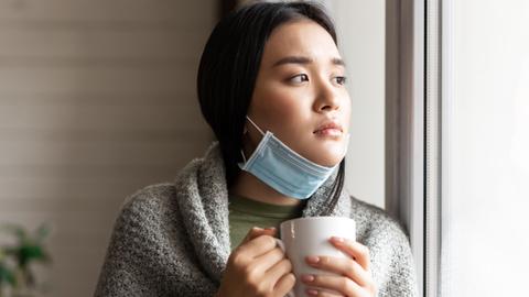 Eine junge asiatische Frau mit heruntergezogenen Mundschutz und einer Tasse in der Hand. 