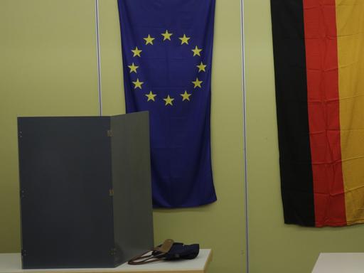 Zu sehen ist eine Wahlkabine in Berlin. Im Hintergrund die Flaggen der EU und der BRD.