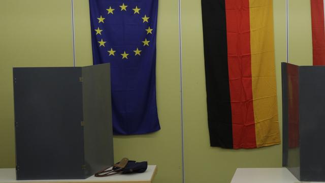 Zu sehen ist eine Wahlkabine in Berlin. Im Hintergrund die Flaggen der EU und der BRD.
