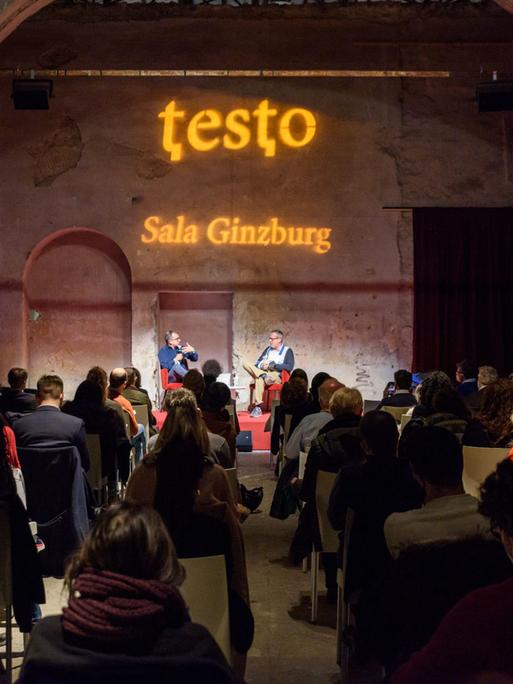 Blick in einen alten Saal während der Buchmesse Testo mit Publikum. Auf der Bühne unterhalten sich zwei Personen über ein Buch.