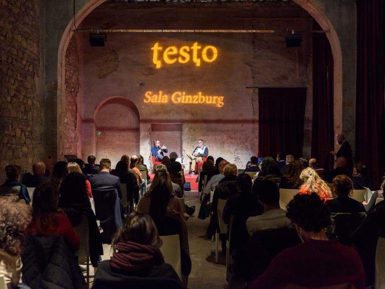 Blick in einen alten Saal während der Buchmesse Testo mit Publikum. Auf der Bühne unterhalten sich zwei Personen über ein Buch.