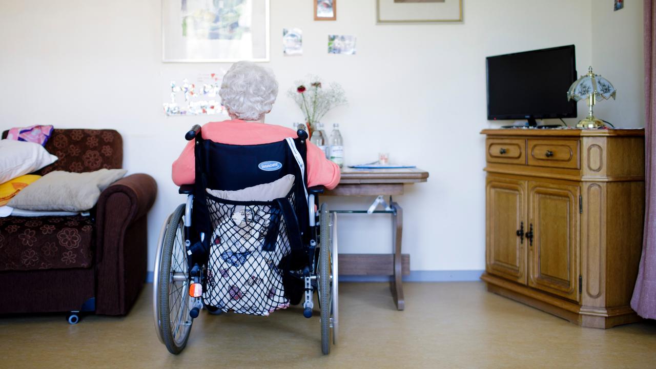 Eine ältere Frau im Rollstuhl sitzt in ihrem Zimmer in einer Pflegeeinrichtung. Sie ist von hinten zu sehen. Auf einer Seite steht ein ein Sessel, auf der anderen ein Schränkchen mit einem Flachbildfernseher darauf. Eine ältere Frau im Rollstuhl sitzt in ihrem Zimmer in einer Pflegeeinrichtung. Sie ist von hinten zu sehen. Auf einer Seite steht ein ein Sessel, auf der anderen ein Schränkchen mit einem Flachbildfernseher darauf.