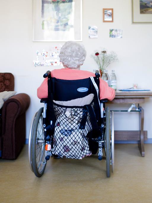 Eine ältere Frau im Rollstuhl sitzt in ihrem Zimmer in einer Pflegeeinrichtung. Sie ist von hinten zu sehen. Auf einer Seite steht ein ein Sessel, auf der anderen ein Schränkchen mit einem Flachbildfernseher darauf. Eine ältere Frau im Rollstuhl sitzt in ihrem Zimmer in einer Pflegeeinrichtung. Sie ist von hinten zu sehen. Auf einer Seite steht ein ein Sessel, auf der anderen ein Schränkchen mit einem Flachbildfernseher darauf.