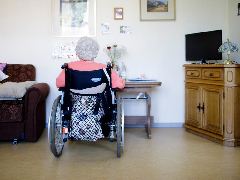 Eine ältere Frau im Rollstuhl sitzt in ihrem Zimmer in einer Pflegeeinrichtung. Sie ist von hinten zu sehen. Auf einer Seite steht ein ein Sessel, auf der anderen ein Schränkchen mit einem Flachbildfernseher darauf. Eine ältere Frau im Rollstuhl sitzt in ihrem Zimmer in einer Pflegeeinrichtung. Sie ist von hinten zu sehen. Auf einer Seite steht ein ein Sessel, auf der anderen ein Schränkchen mit einem Flachbildfernseher darauf.