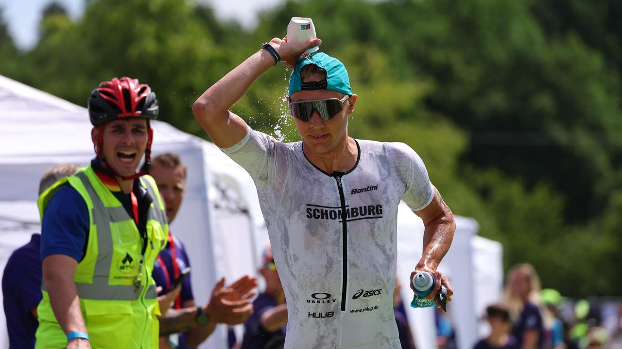 Der deutsche Triathlet Jonas Schomburg schüttet sich beim Laufen Wasser über den Kopf. Neben ihm steht ein Helfer und feuert ihn an.