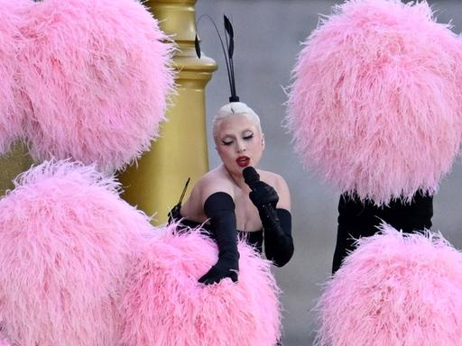 Sängerin Lady Gaga vor einer goldenen Treppe, umgeben von riesigen rosa Pompons.