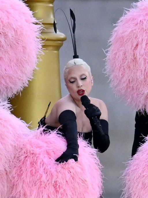 Sängerin Lady Gaga vor einer goldenen Treppe, umgeben von riesigen rosa Pompons.
