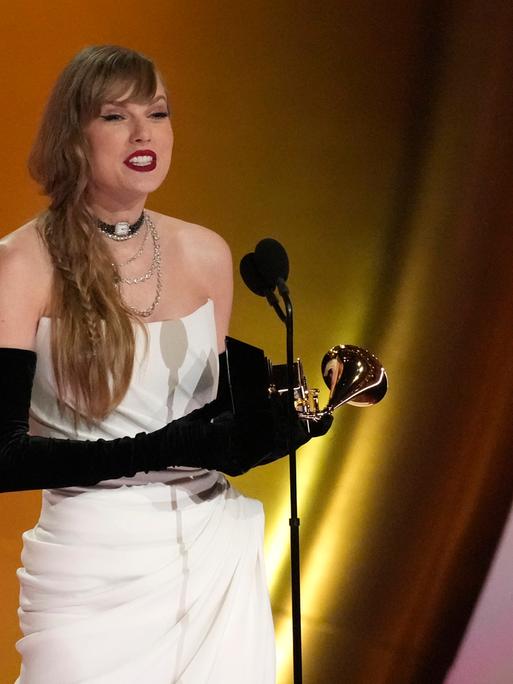 Taylor Swift steht auf der Bühne und hält eine Dankesrede. In der Hand hält sie einen Grammy. Sie trägt ein schulterfreies Kleid und lange Abendhandschuhe.