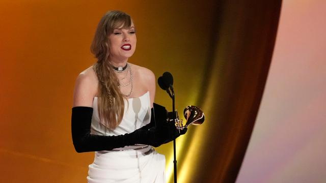 Taylor Swift steht auf der Bühne und hält eine Dankesrede. In der Hand hält sie einen Grammy. Sie trägt ein schulterfreies Kleid und lange Abendhandschuhe.