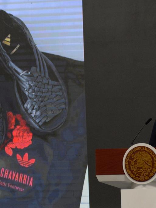 Mexikos Präsidentin Claudia Sheinbaum steht an einem Rednerpult. Hinter ihr sind auf einer Leinwand ein schwarzes Paar der umstrittenen von Adidas designten Huarache-Sandalen zu sehen. 