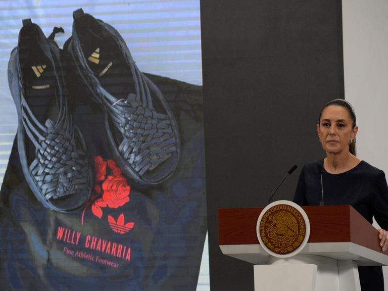 Mexikos Präsidentin Claudia Sheinbaum steht an einem Rednerpult. Hinter ihr sind auf einer Leinwand ein schwarzes Paar der umstrittenen von Adidas designten Huarache-Sandalen zu sehen. 
