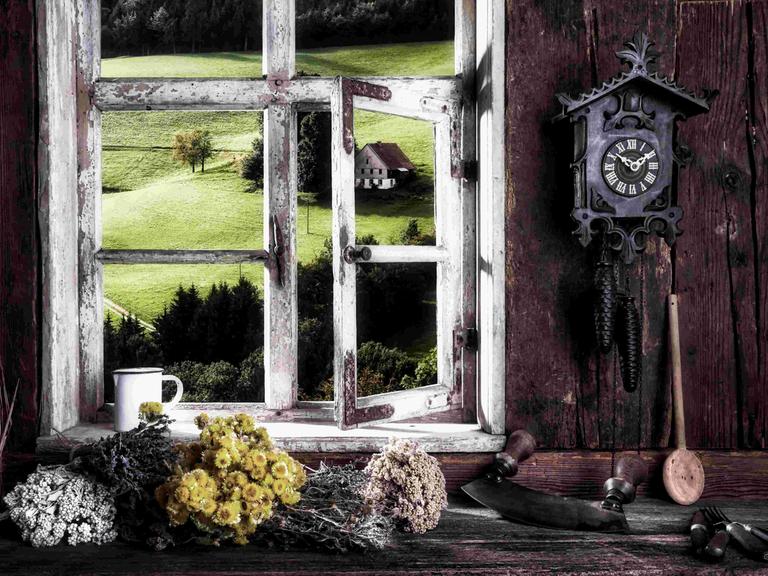 Blick aus dem Fenster einer hölzernen Bauernstube auf eine hügelige Weidelandschaft, in der Stube hängt eine Kuckucksuhr.