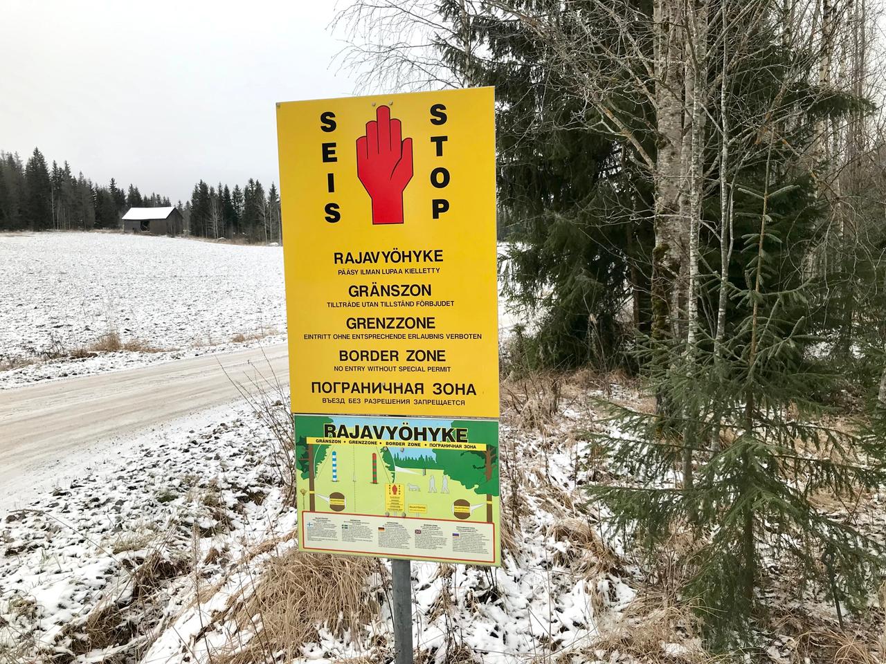 Ein gelbes Schild weist in mehreren Sprachen auf den Grenzbereich zwischen Finnland und Russland hin. Es steht entlang eines verschneiten Weges am Waldrand. Im Hintergrund ist ein Haus zu sehen.