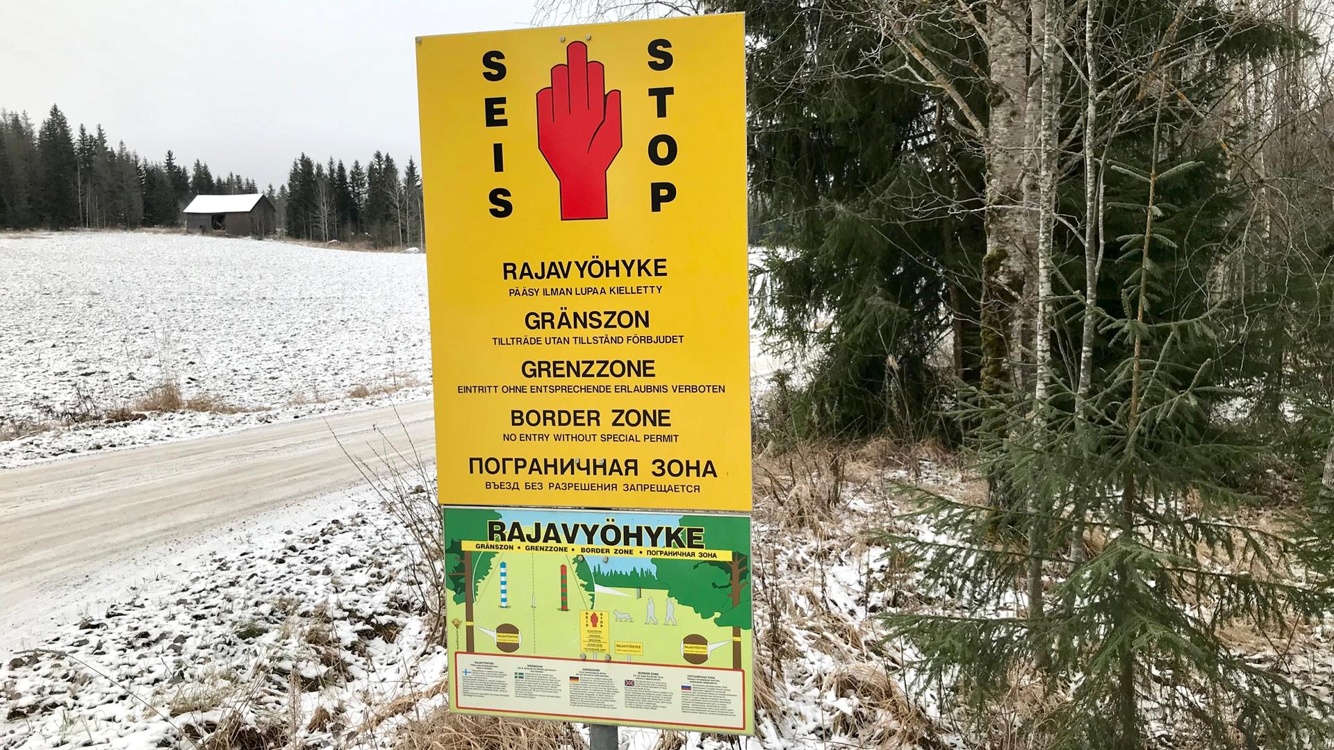 Ein gelbes Schild weist in mehreren Sprachen auf den Grenzbereich zwischen Finnland und Russland hin. Es steht entlang eines verschneiten Weges am Waldrand. Im Hintergrund ist ein Haus zu sehen.