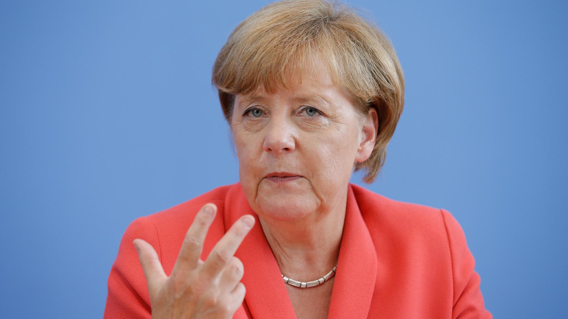 Angela Merkel sitzt vor blauem Hintergrund. Sie trägt eine rötliche Jacke und eine Kette um den Hals. Angela Merkel sitzt vor blauem Hintergrund. Sie trägt eine rötliche Jacke und eine Kette um den Hals.