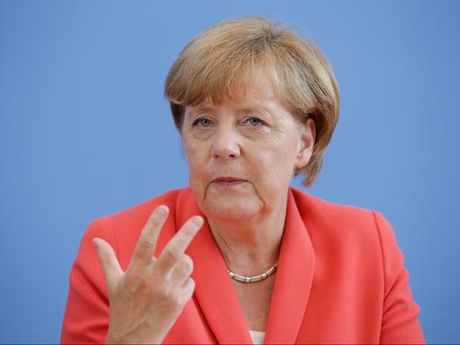 Angela Merkel sitzt vor blauem Hintergrund. Sie trägt eine rötliche Jacke und eine Kette um den Hals.