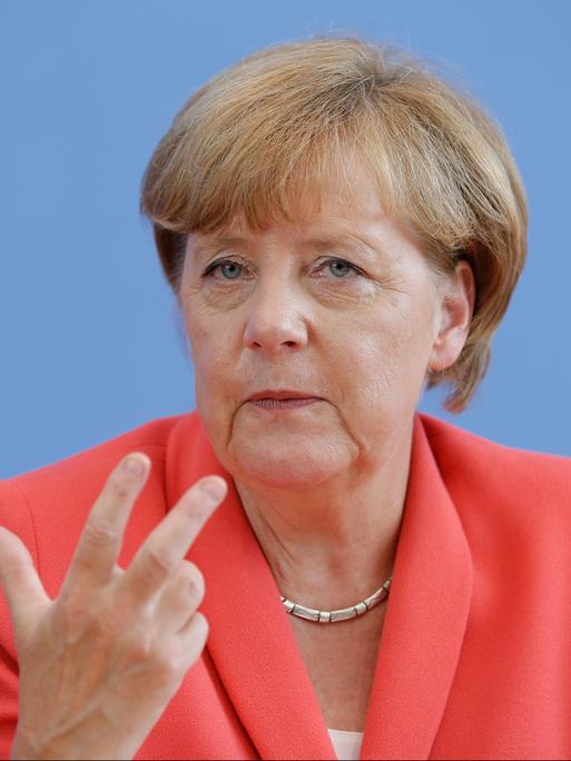 Angela Merkel sitzt vor blauem Hintergrund. Sie trägt eine rötliche Jacke und eine Kette um den Hals.
