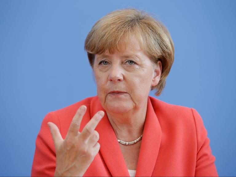 Angela Merkel sitzt vor blauem Hintergrund. Sie trägt eine rötliche Jacke und eine Kette um den Hals.