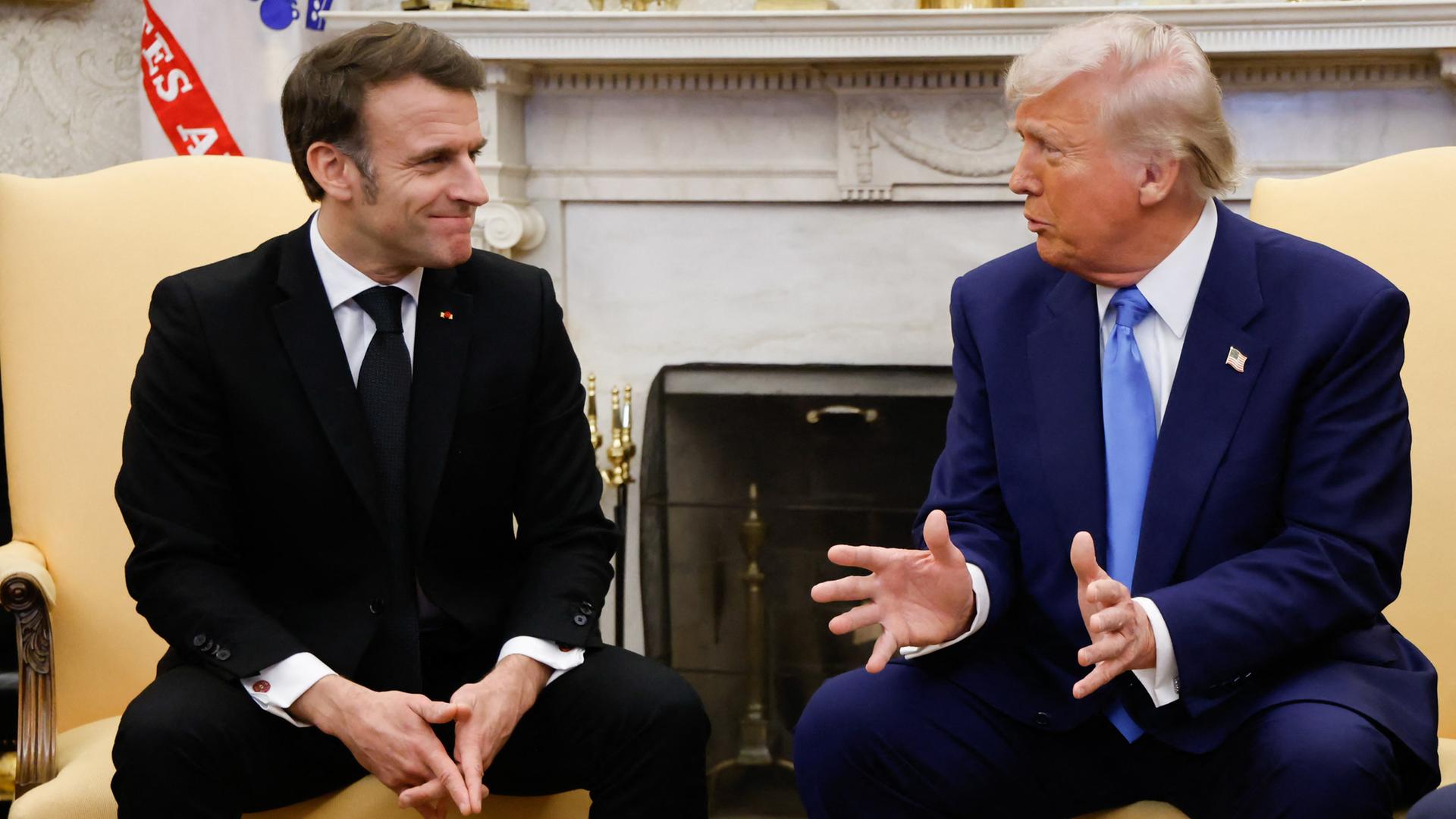 Macron und Trump sitzen nebeneinander und schauen sich an. Sie führen ein Gespräch.