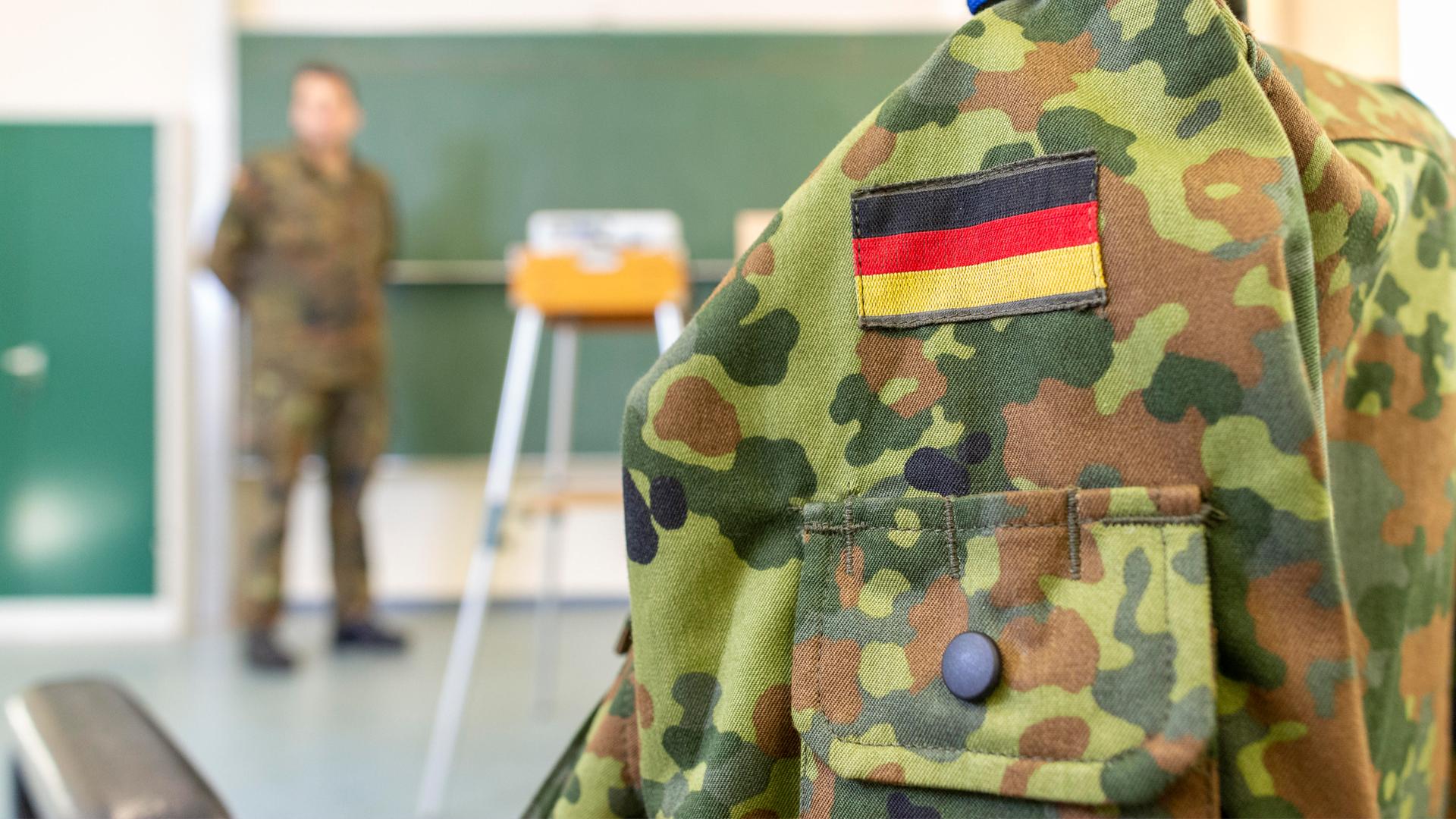 Eine Person in Bundeswehruniform steht an einer Tafel in einem Klassenraum. Im Vordergrund ist eine Bundeswehruniform auf einer Kleiderpuppe zu sehen.