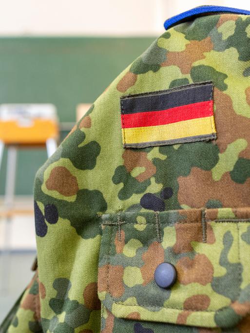 Eine Person in Bundeswehruniform steht an einer Tafel in einem Klassenraum. Im Vordergrund ist eine Bundeswehruniform auf einer Kleiderpuppe zu sehen. Eine Person in Bundeswehruniform steht an einer Tafel in einem Klassenraum. Im Vordergrund ist eine Bundeswehruniform auf einer Kleiderpuppe zu sehen.