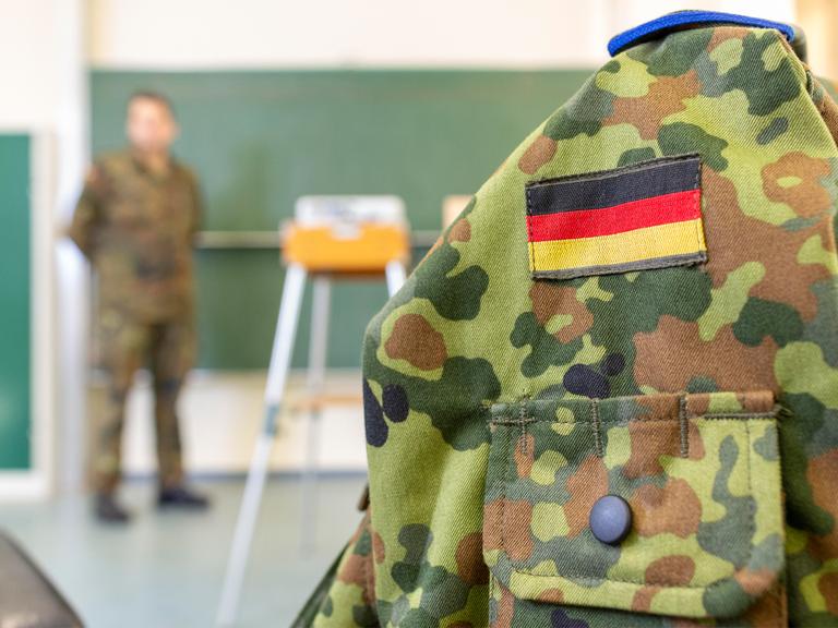 Eine Person in Bundeswehruniform steht an einer Tafel in einem Klassenraum. Im Vordergrund ist eine Bundeswehruniform auf einer Kleiderpuppe zu sehen. Eine Person in Bundeswehruniform steht an einer Tafel in einem Klassenraum. Im Vordergrund ist eine Bundeswehruniform auf einer Kleiderpuppe zu sehen.