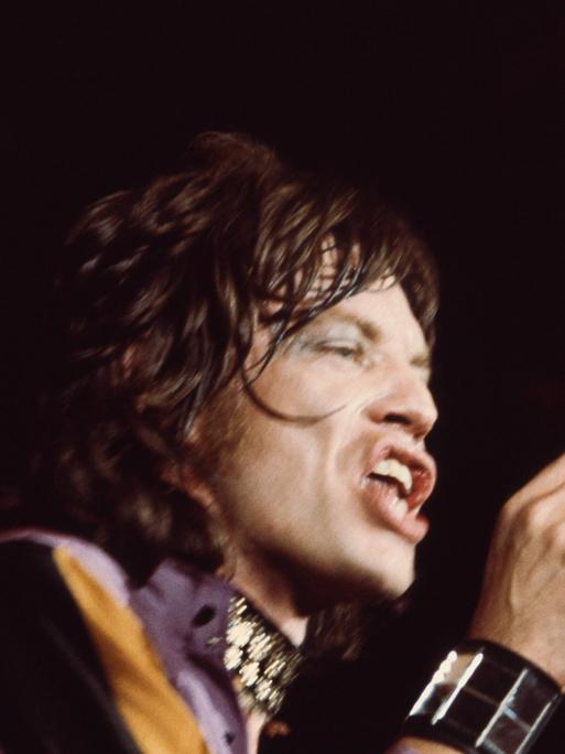 Mick Jagger singt vor schwarzem Hintergrund in ein Mikrofon