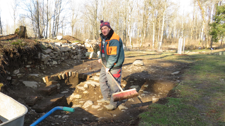 Helmut Roith fegt an der Grabungsstelle mit einem Besen die Mauerreste von Erde frei