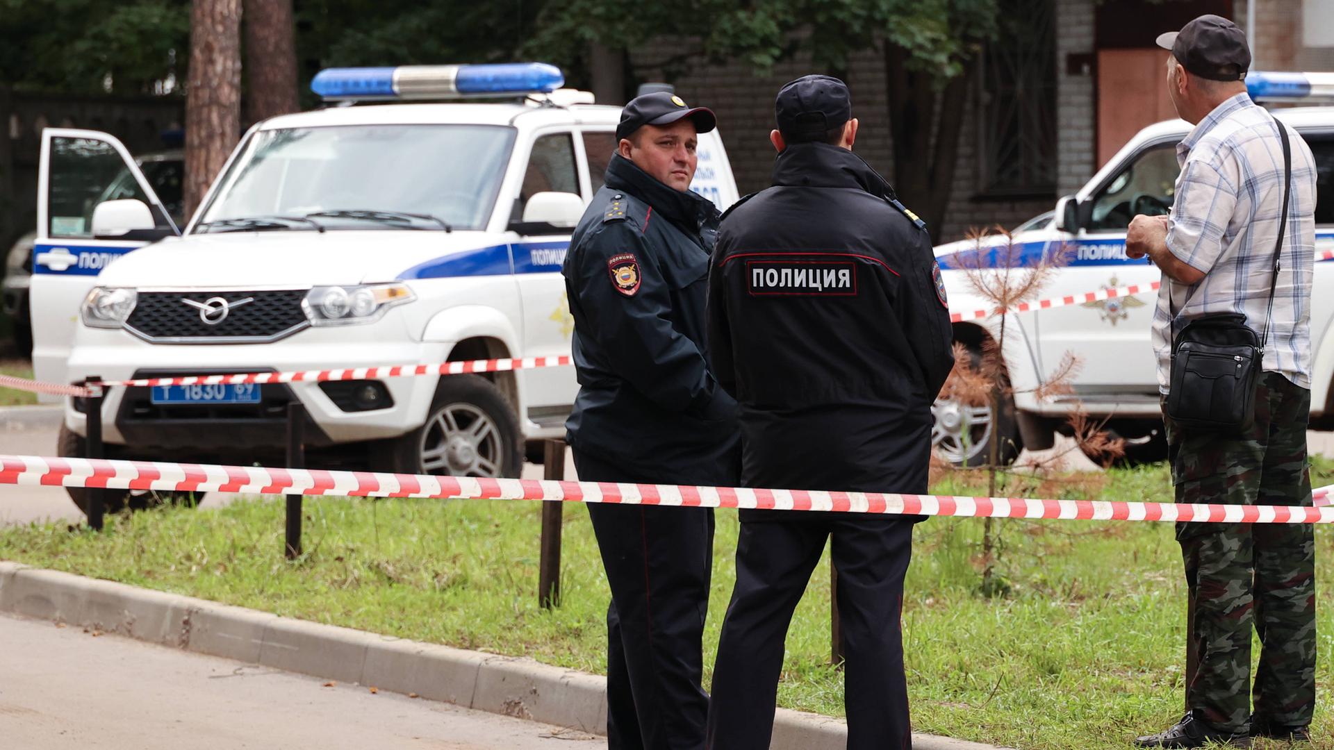 Zwei russische Polizisten stehen in Uniform mit dem Rücken zur Kamera. Um sie herum Absperrungsband, hinter ihnen steht ein Polizeiauto.