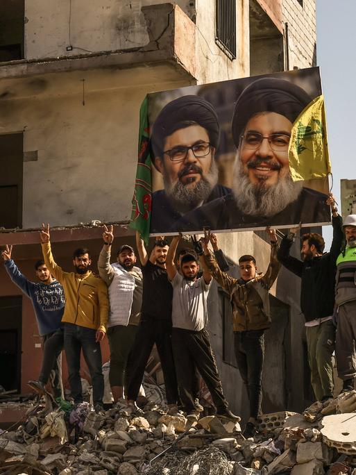 Menschen in dem von der Hisbollah kontrollierten Dahieh Viertel im Süden Beiruts in Trümmern halten ein Foto eines Politikers mit schwarzem Turban  hoch