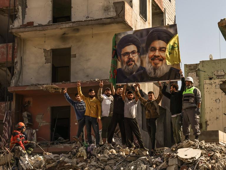 Menschen in dem von der Hisbollah kontrollierten Dahieh Viertel im Süden Beiruts in Trümmern halten ein Foto eines Politikers mit schwarzem Turban  hoch