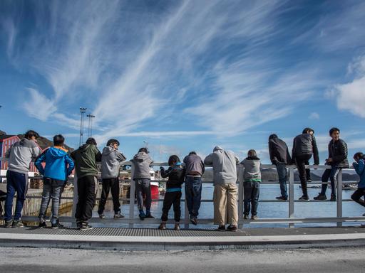 Eine Gruppe jugendlicher Jungs steht an einer hölzernen Uferbegrenzung. Manche sind daran gelehnt und schauen aufs Wasser, andere sitzen rittlings darauf.