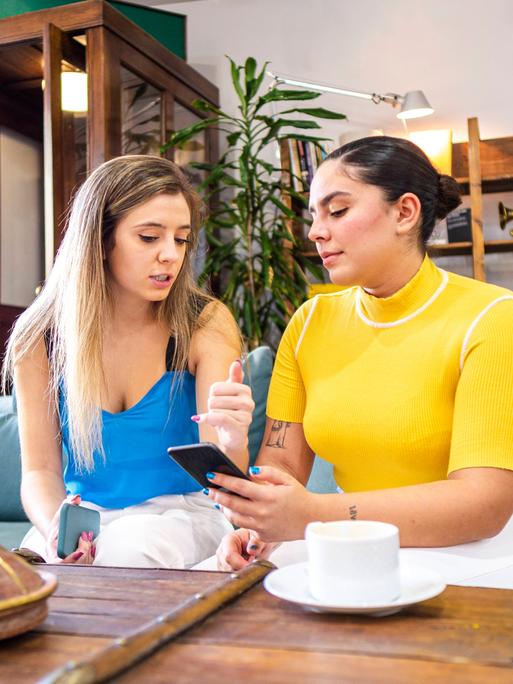 Zwei Frauen sitzen nebeneinander auf einer Couch und schauen gemeinsam auf ein Handy. Dabei diskutieren sind miteinadner. Zwei Frauen sitzen nebeneinander auf einer Couch und schauen gemeinsam auf ein Handy. Dabei diskutieren sind miteinadner.