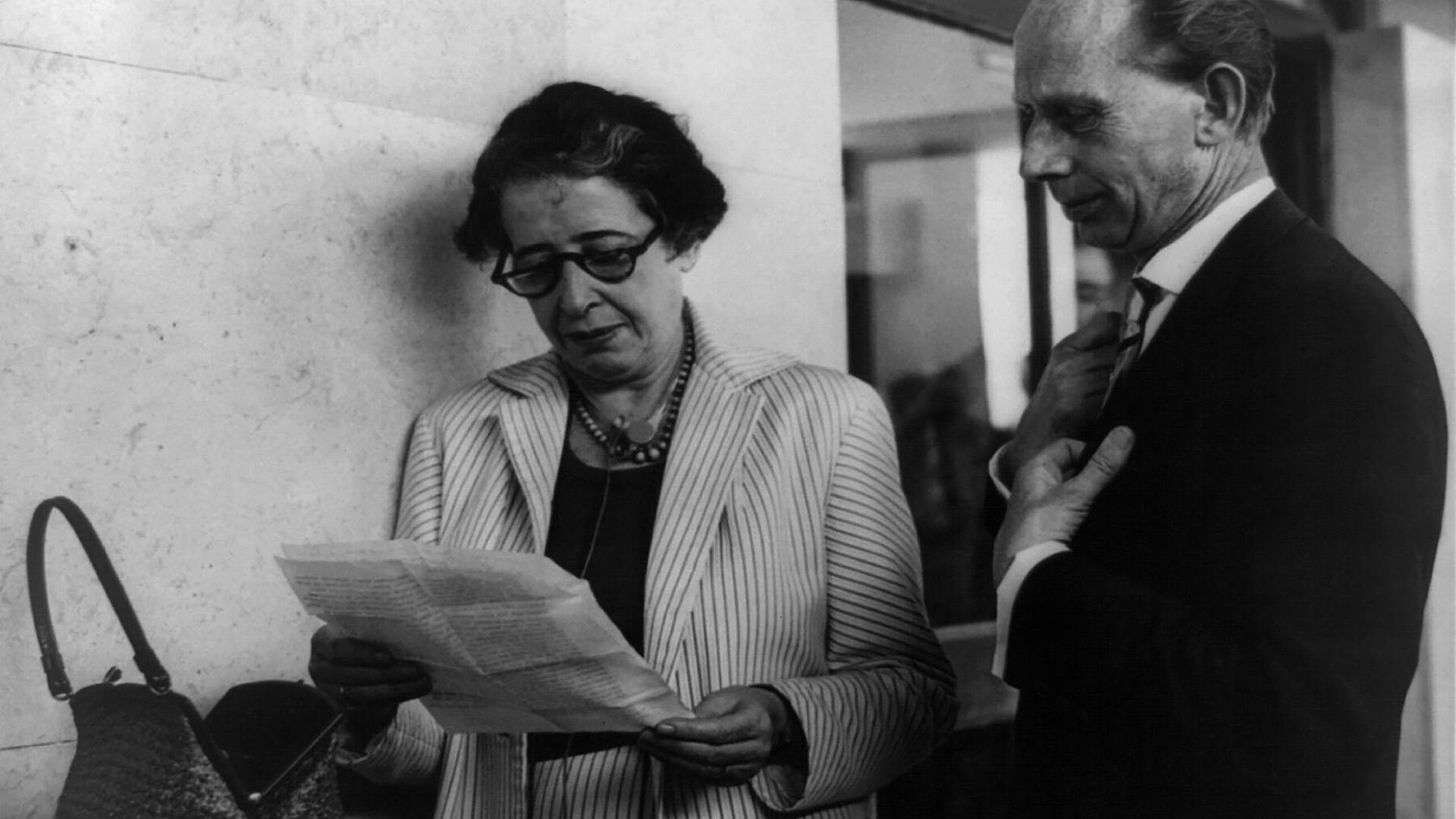 Ein Schwarz-weiß-Foto von circa 1960 zeigt Hannah Arendt, die mit Brille auf ein Blatt Papier schaut, neben ihr ein Reporter-Kollege