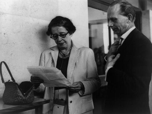 Ein Schwarz-weiß-Foto von circa 1960 zeigt Hannah Arendt, die mit Brille auf ein Blatt Papier schaut, neben ihr ein Reporter-Kollege