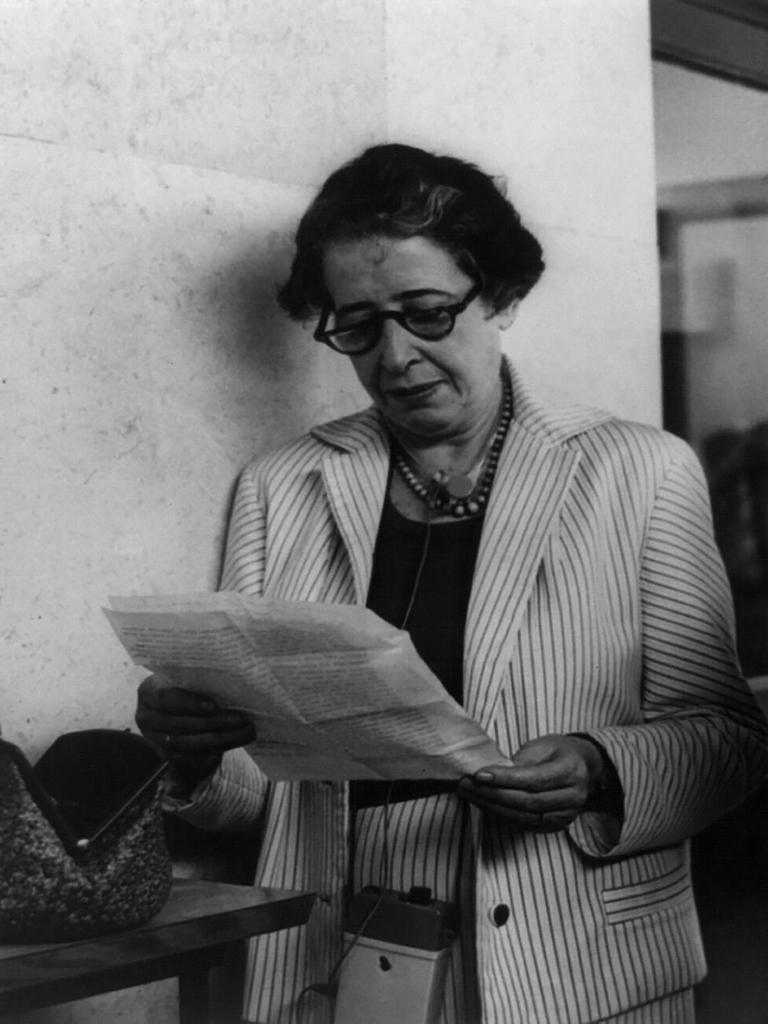 Ein Schwarz-weiß-Foto von circa 1960 zeigt Hannah Arendt, die mit Brille auf ein Blatt Papier schaut, neben ihr ein Reporter-Kollege