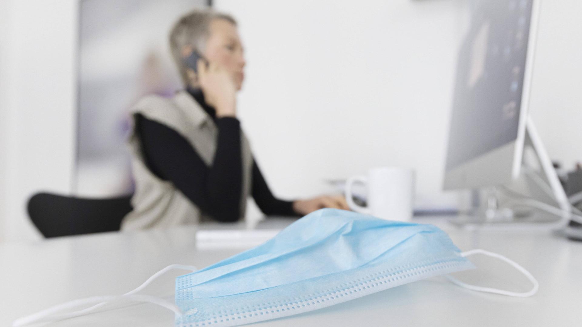 Eine Frau am Arbeitsplatz, die Coronaschutz-Maske liegt daneben.