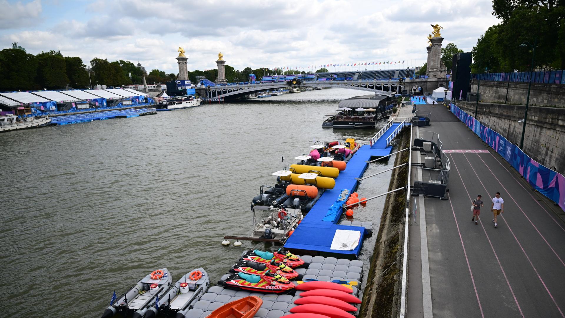 Im Vordergrund eine Straße entlang des Flusses. Am Ufer eine Plattform mit Paddelbooten. im Hintergrudn eine Seine-Brücke. Im Vordergrund eine Straße entlang des Flusses. Am Ufer eine Plattform mit Paddelbooten. im Hintergrudn eine Seine-Brücke.