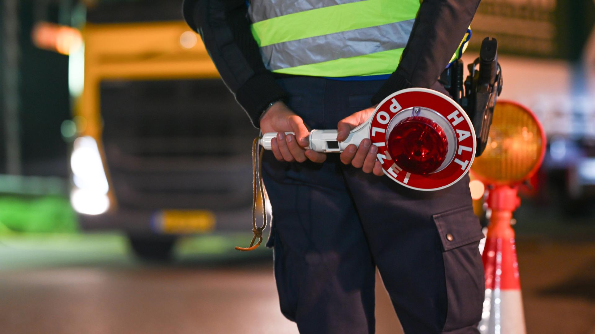 Eine Polizistin oder ein Polizist steht an einer Grenze. Im Hintergrund ist ein Lastwagen mit einem gelben Kennzeichen zu sehen.