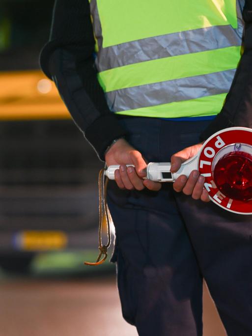 Eine Polizistin oder ein Polizist steht an einer Grenze. Im Hintergrund ist ein Lastwagen mit einem gelben Kennzeichen zu sehen.