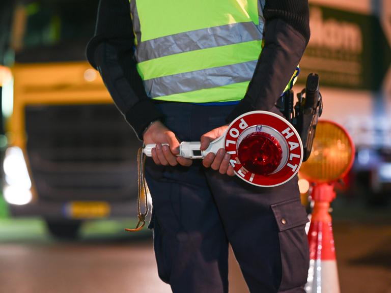 Eine Polizistin oder ein Polizist steht an einer Grenze. Im Hintergrund ist ein Lastwagen mit einem gelben Kennzeichen zu sehen.