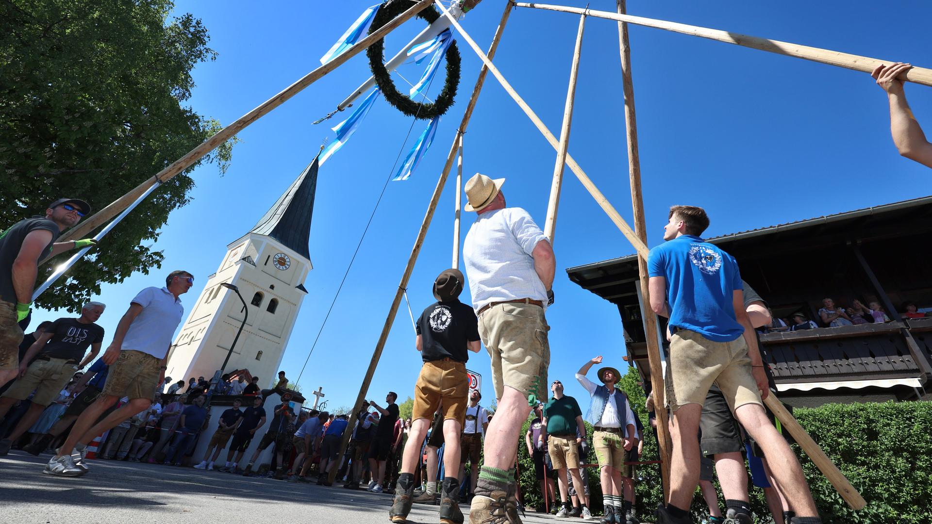 Mehrere Männer richten mit Muskelkraft und der Hilfe von Stangen vor strahlend blauem Himmel den Maibaum auf. Im Hintergrund ist die örtliche Kirche zu sehen. Mehrere Männer richten mit Muskelkraft und der Hilfe von Stangen vor strahlend blauem Himmel den Maibaum auf. Im Hintergrund ist die örtliche Kirche zu sehen.