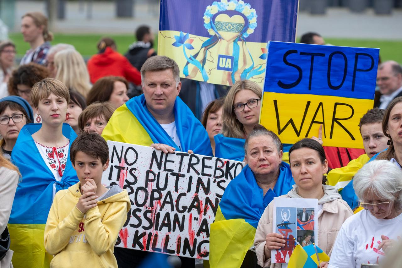 Solidaritätsdemonstration für die Ukraine und gegen einen Auftritt der russischen Sopranistin Anna Netrebko in Wiesbaden. Männer, Frauen und Kinder blicken in Richtung der Kamera. Einige sind in blau-gelbe ukrainische Flaggen gehüllt. Auf einem Schild steht "Stop war", auf Deutsch "Beendet Krieg".
