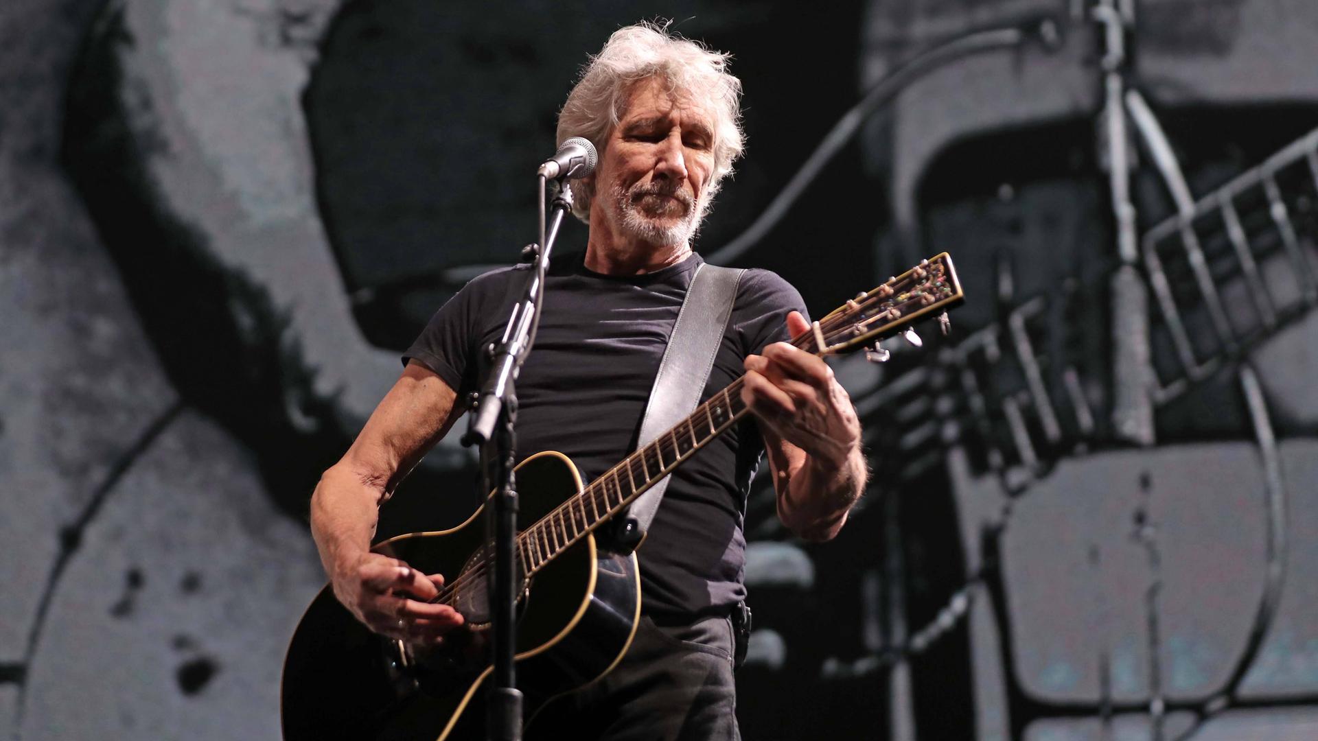 Der Pink Floyd-Mitgründer Roger Waters 2020 bei einem Konzert in Mexiko: Er steht auf einer abgedunkelten Bühne hinter einem Mikrofon und spielt eine akustische Gitarre.