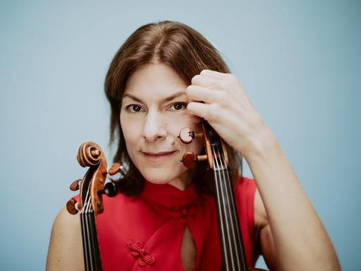 Nahaufnahme einer Frau mit braunen Haaren und rotem Top vor hellblauem Hintergrund. Sie blickt nachdenklich und direkt in die Kamera. In der linken Hand und im rechten Arm hält sie eine Geige und eine Bratsche.