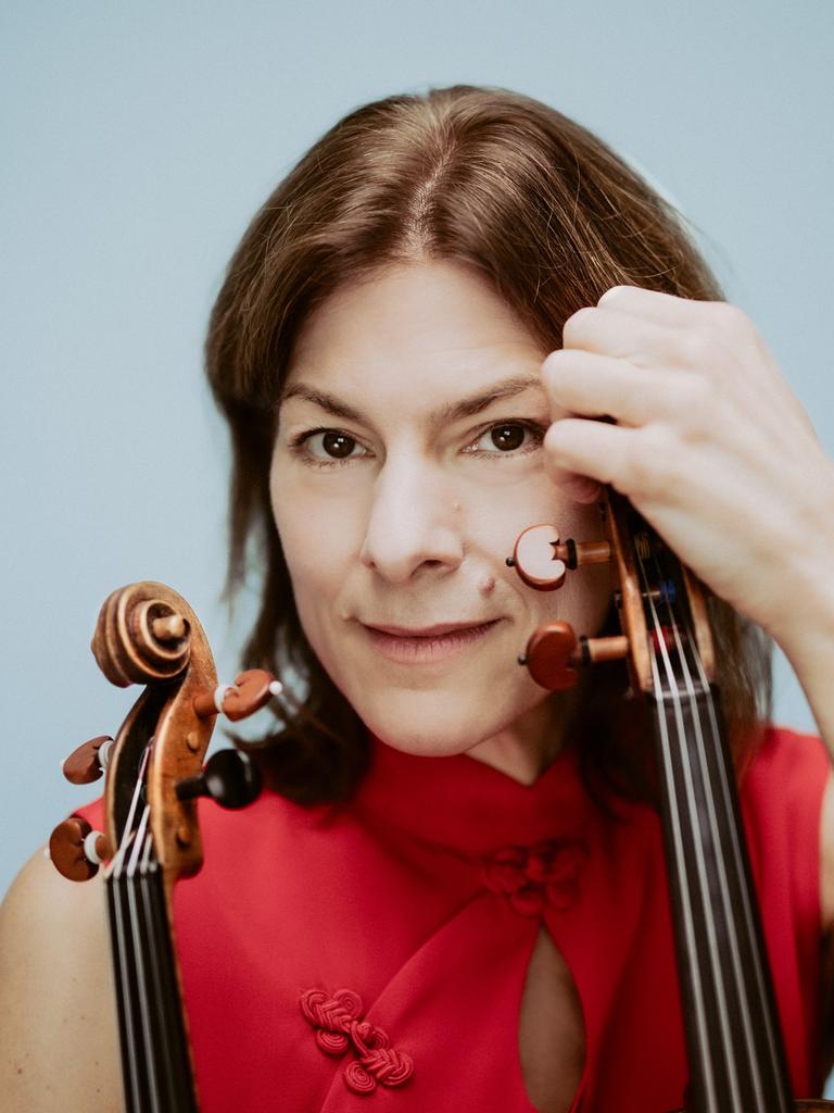 Nahaufnahme einer Frau mit braunen Haaren und rotem Top vor hellblauem Hintergrund. Sie blickt nachdenklich und direkt in die Kamera. In der linken Hand und im rechten Arm hält sie eine Geige und eine Bratsche.