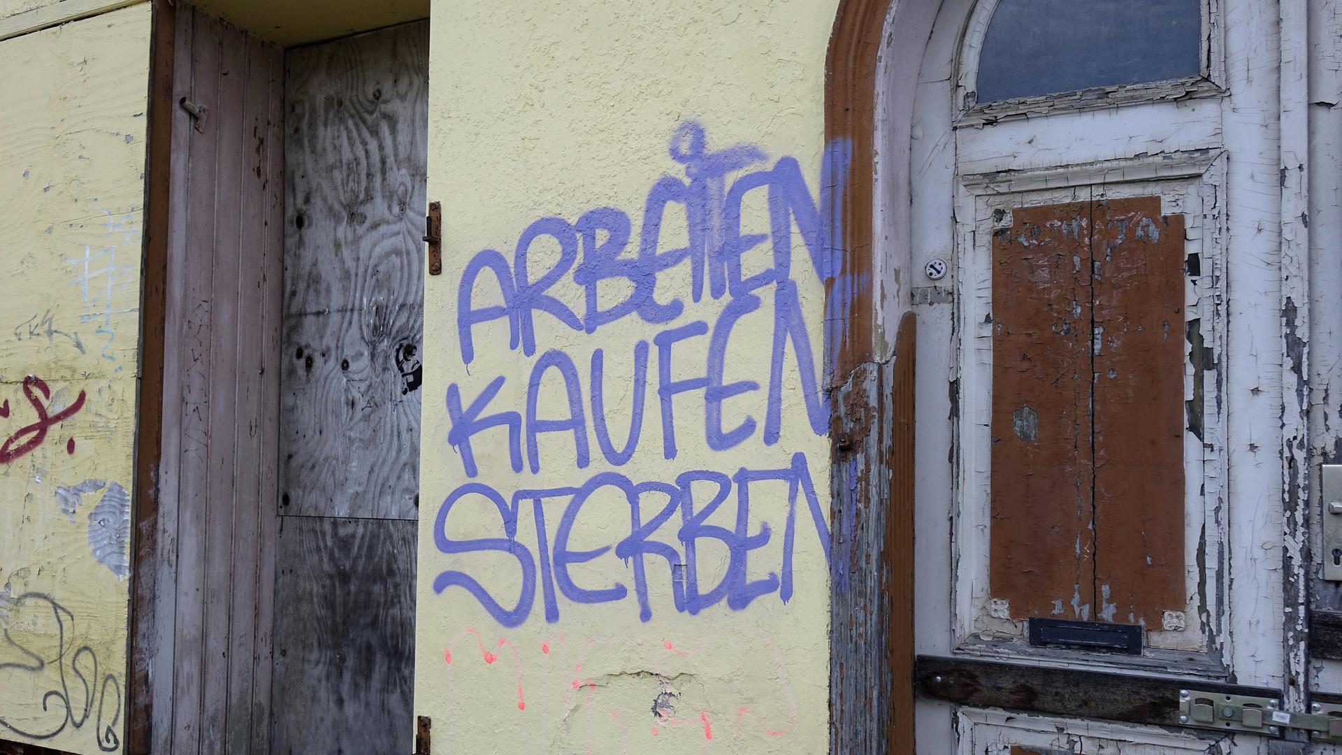 Ein Graffito an einer Hauswand. "Arbeiten", "Kaufen" und "Sterben" stehen in violetter Schrift untereinander an einer Hauswand. Rechts und links der gelblichen Mauer ist Holz zu sehen, vermutlich von einer Tür. 
