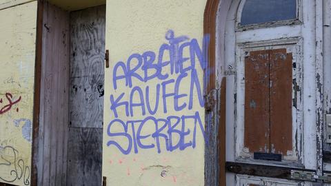 Ein Graffito an einer Hauswand. "Arbeiten", "Kaufen" und "Sterben" stehen in violetter Schrift untereinander an einer Hauswand. Rechts und links der gelblichen Mauer ist Holz zu sehen, vermutlich von einer Tür. 