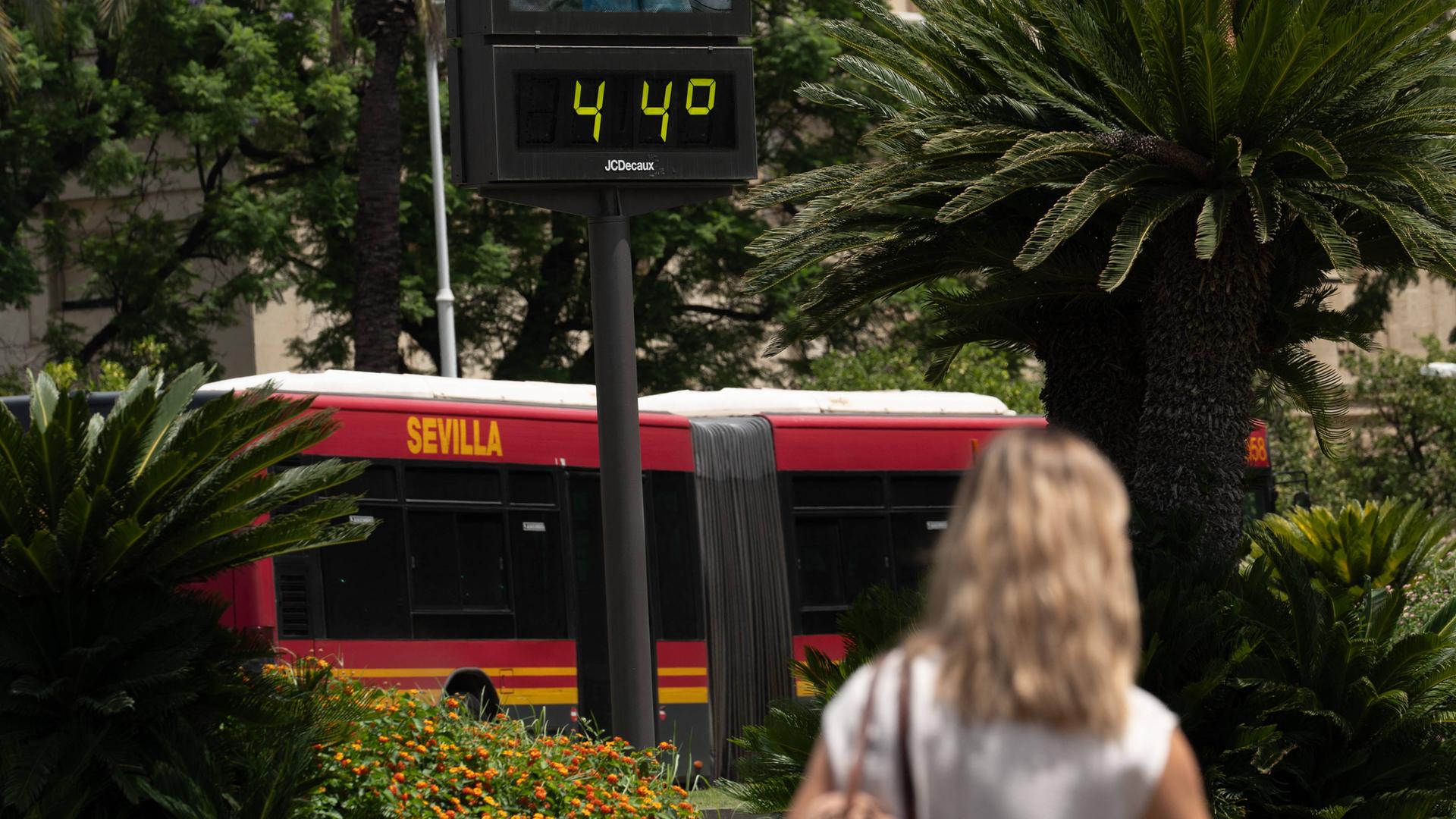 Eine Frau läuft neben einem Bus vorbei auf dem das Wort Sevilla steht. Über ihrem Kopf zeigt ein Thermometer die Temperatur von 44 Grad Celsius an.